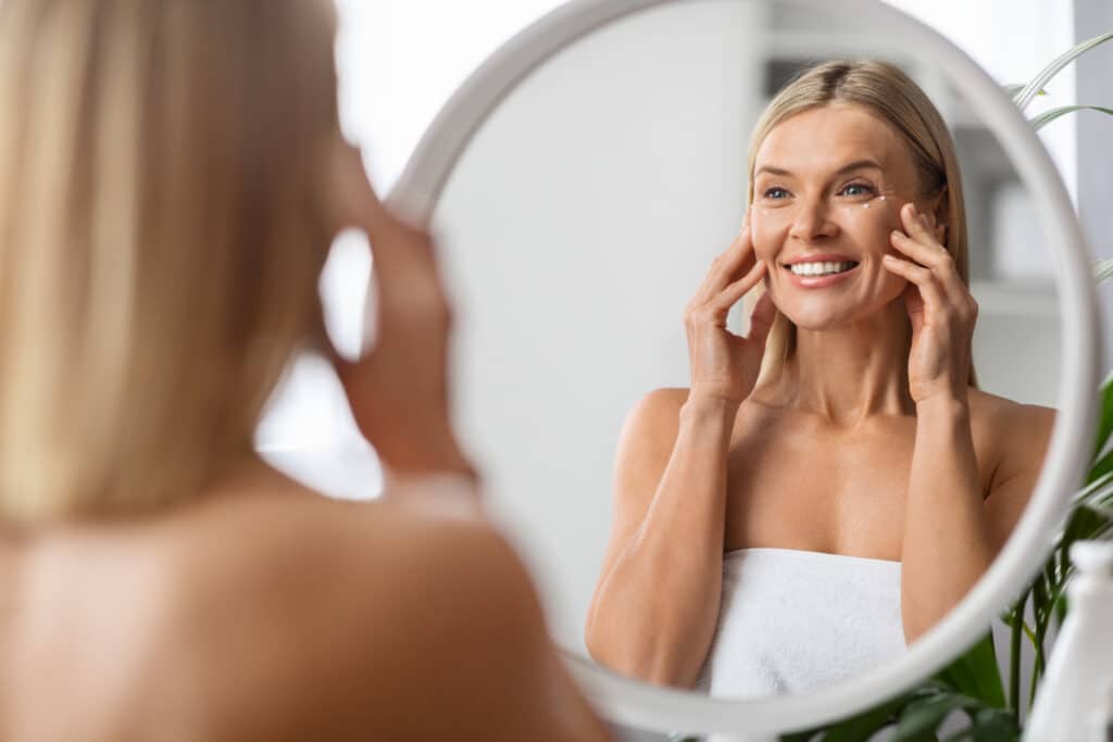 Anti-Aging Cosmetics. Beautiful Middle Aged Woman Applying Eye Cream While Standing Near Mirror At Home, Attractive Mature Lady Touching Her Face, Enjoying Daily Beauty Routine, Selective Focus