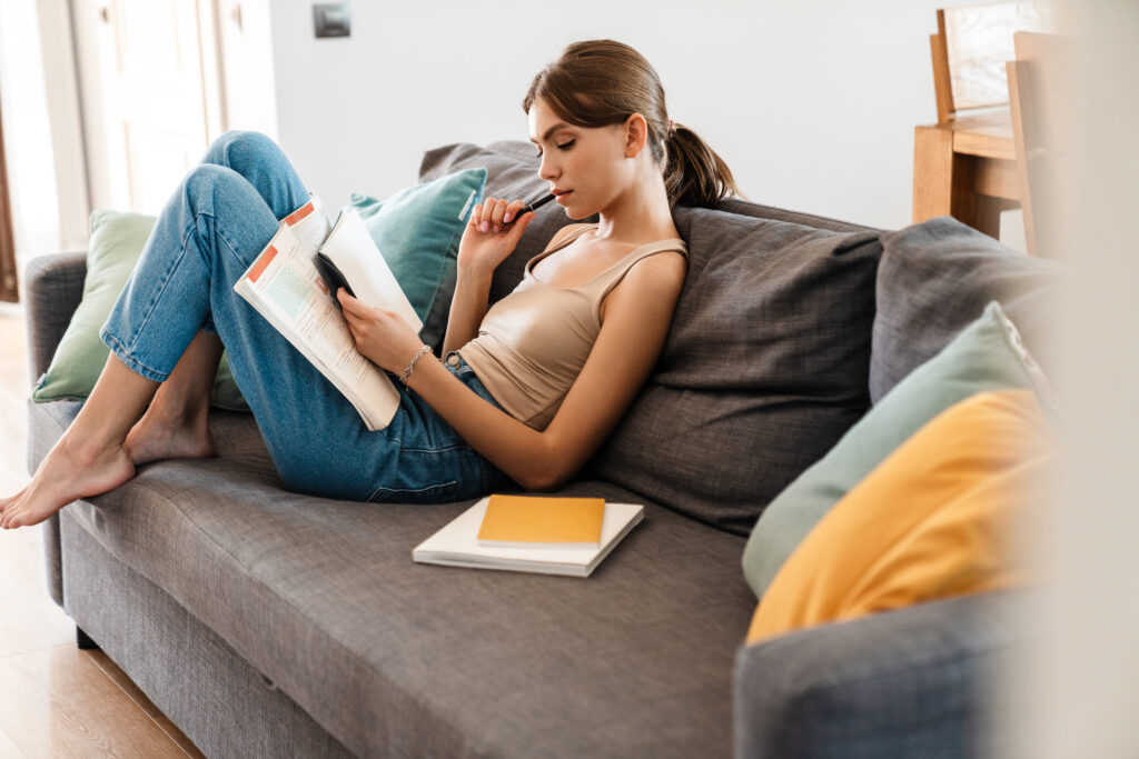 Attractive young woman studying while sitting on couch at home, taking notes
