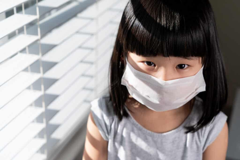 Little chinese girl wearing mask imprison at home standing next to window for corona virus or air pollution protection concept