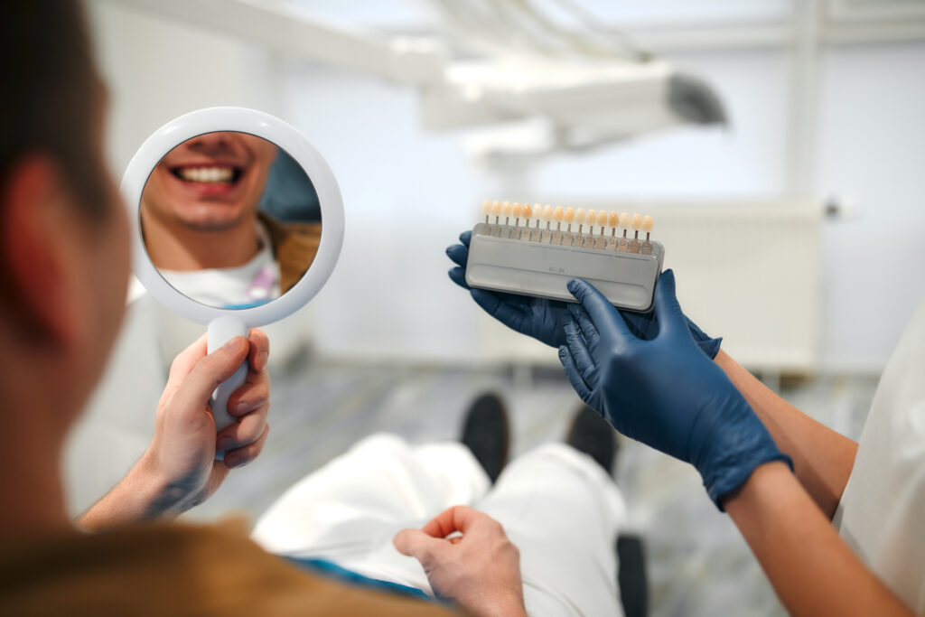 Medicine, dentistry and healthcare concept - closeup of a dentist with tooth color samples choosing a shade for a male patient's teeth in a dental clinic looking at a mirror.