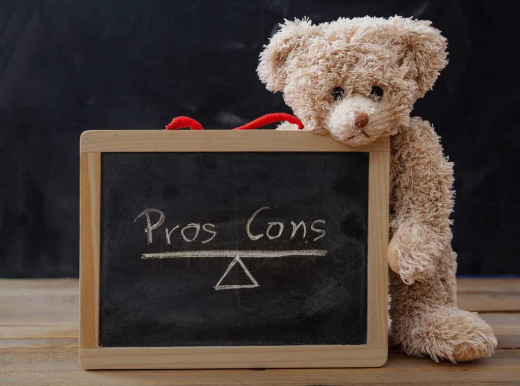 Pros contra cons concept. Teddy bear on wooden table holding a framed chalkboard with empty list on, blackboard background, for analysis and decision making at school, business.