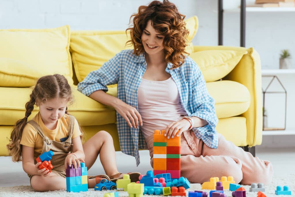 smiling nanny and adorable kid playing with multicolored building blocks on floor