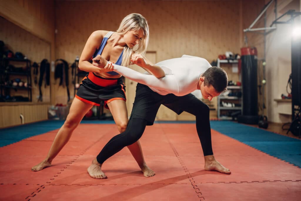 Woman makes elbow kick, self-defense workout with male personal trainer, gym interior on background. Female person on training, self defense practice