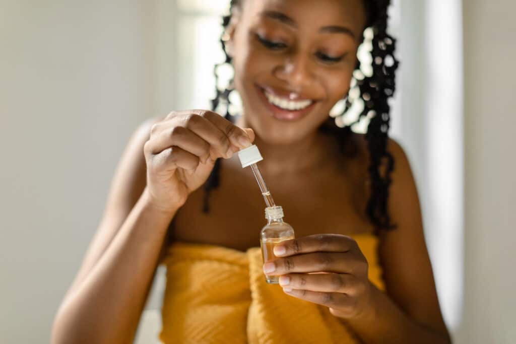 Black lady holding open bottle with moisturizing face serum, sitting in bedroom and smiling, enjoying making daily skin care treatments, selective focus, closeup
