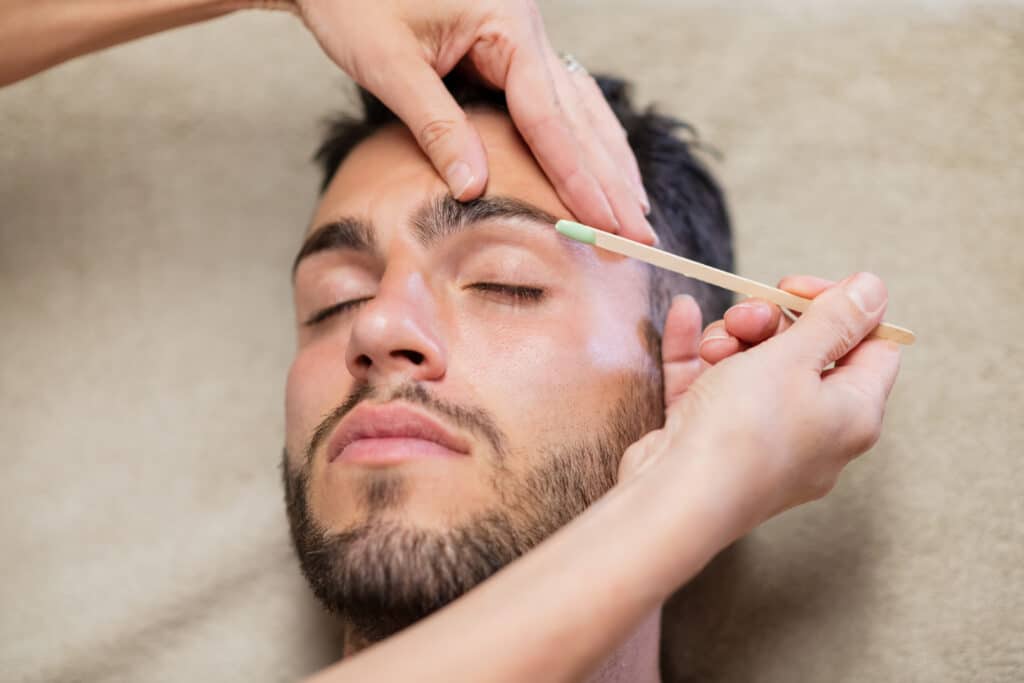 using stick to apply wax on eyebrow of a bearded young man with closed eyes during work in beauty studio