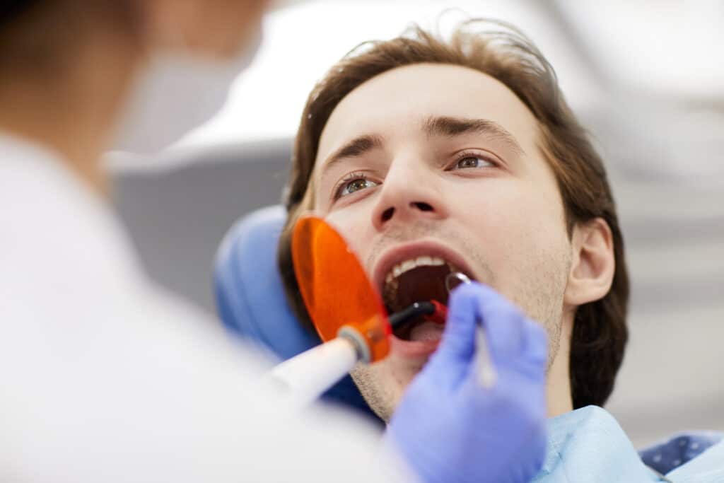 Dentist performing laser teeth whitening on man sitting in dental chair with mouth open