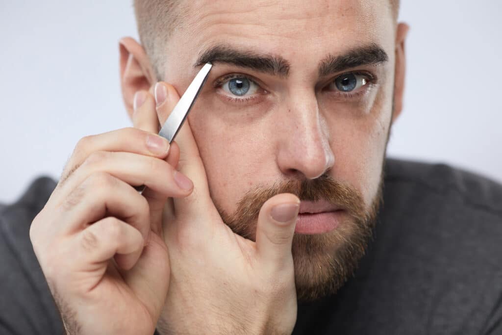 Horizontal close up portrait of handsome young man tweezing his eyebrows looking at camera