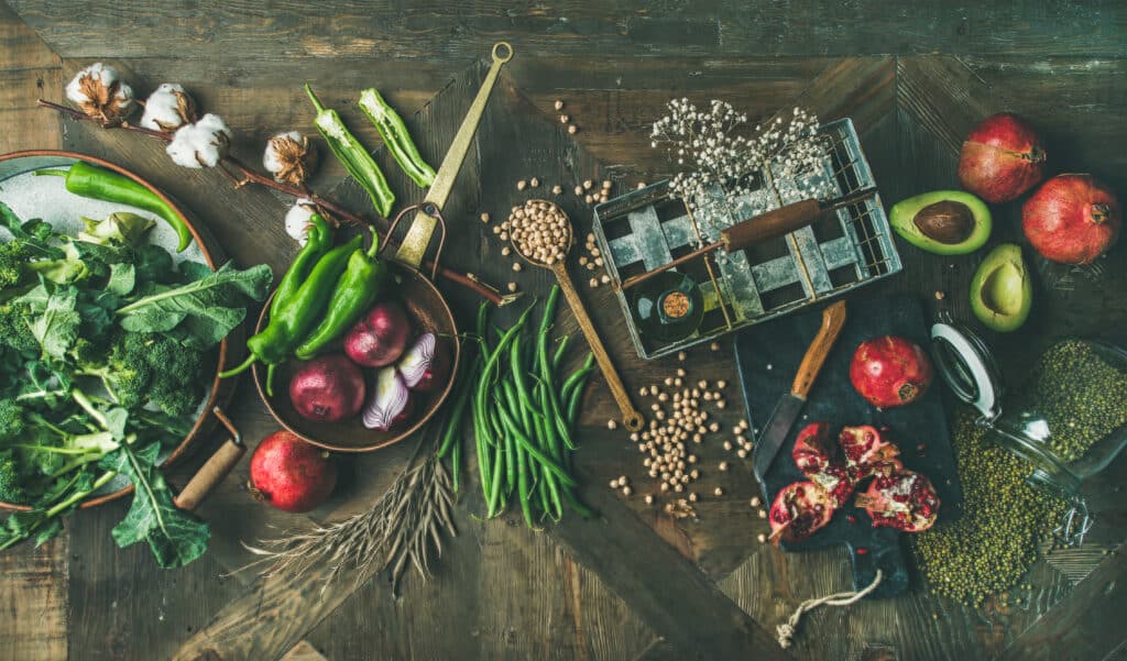 Winter vegetarian or vegan food cooking ingredients. Flat-lay of seasonal vegetables and fruits, beans, cereals, kitchen utencils, dried flowers, olive oil over wooden background, top view