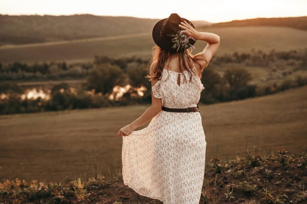 Back view of unrecognizable carefree female in white summer dress and sunhat standing in meadow and enjoying evening