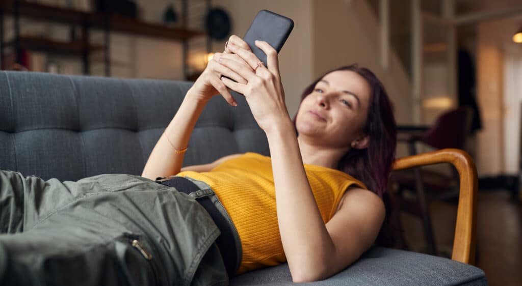 Young Woman Lying On Sofa At Home Looking At Mobile Phone Messages And Social Media